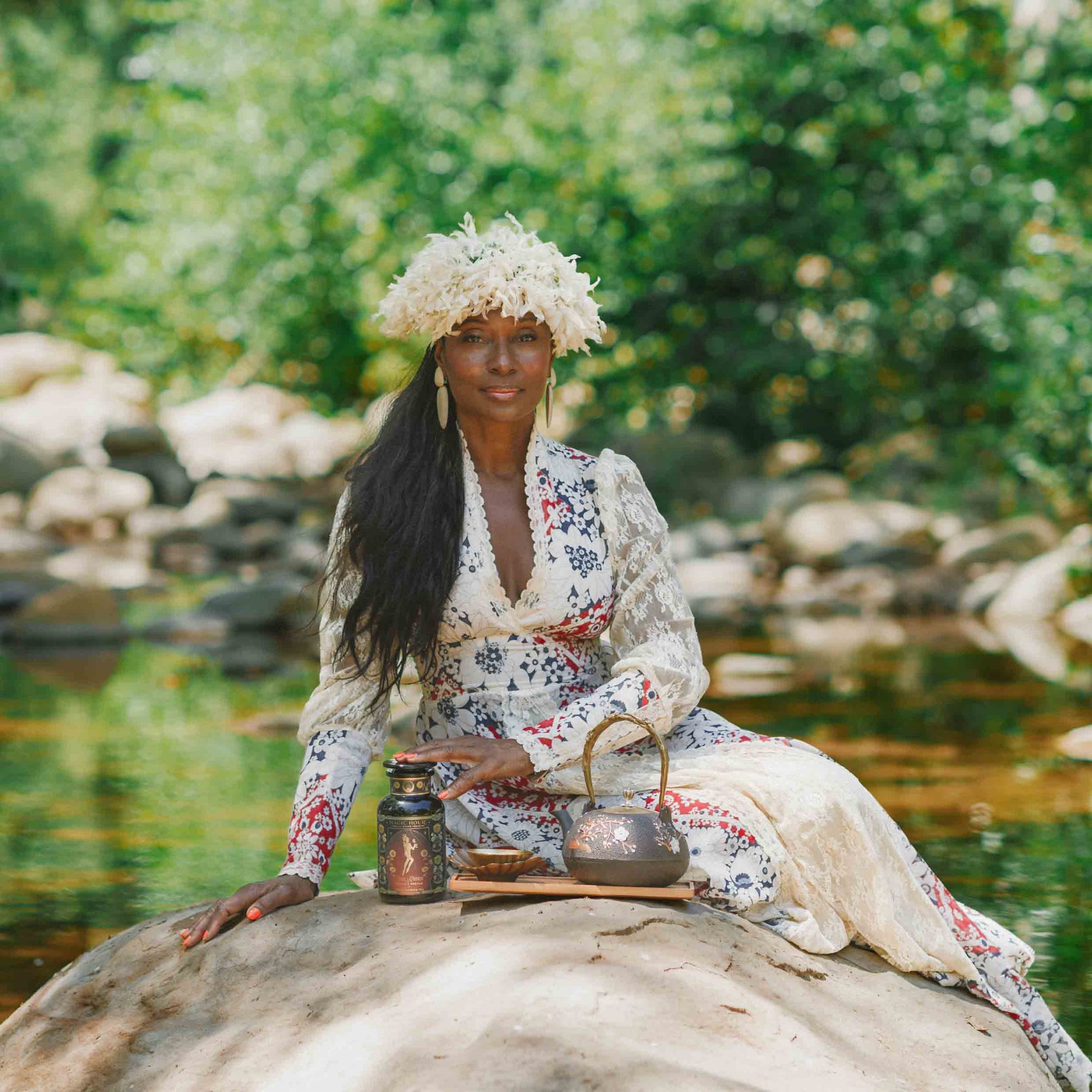 Clad in a floral dress and hat, a woman sits serenely on a rock beside a creek, enveloped by lush greenery. Channeling the grace of a goddess archetype, she savors a tranquil outdoor moment with her teapot, cup, and jar at her side—bringing to life the gentle ritual suggested by the Monthly Magic Tea Journey Subscription as she enjoys afternoon tea embraced by nature.