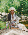 Clad in a floral dress and hat, a woman sits serenely on a rock beside a creek, enveloped by lush greenery. Channeling the grace of a goddess archetype, she savors a tranquil outdoor moment with her teapot, cup, and jar at her side—bringing to life the gentle ritual suggested by the Monthly Magic Tea Journey Subscription as she enjoys afternoon tea embraced by nature.
