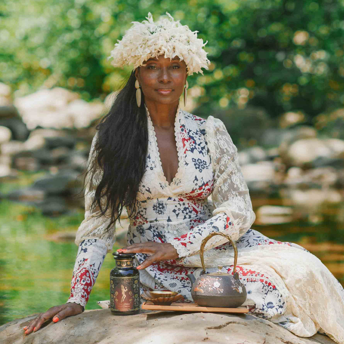 A woman in a long, patterned dress and flower hat sits on a rock by a stream, accompanied by a teapot, cup, and a jar of Oshun’s Nectar: Honeyed Herbal Tea for Joy & Radiance. Green trees and flowing water create the tranquil background.