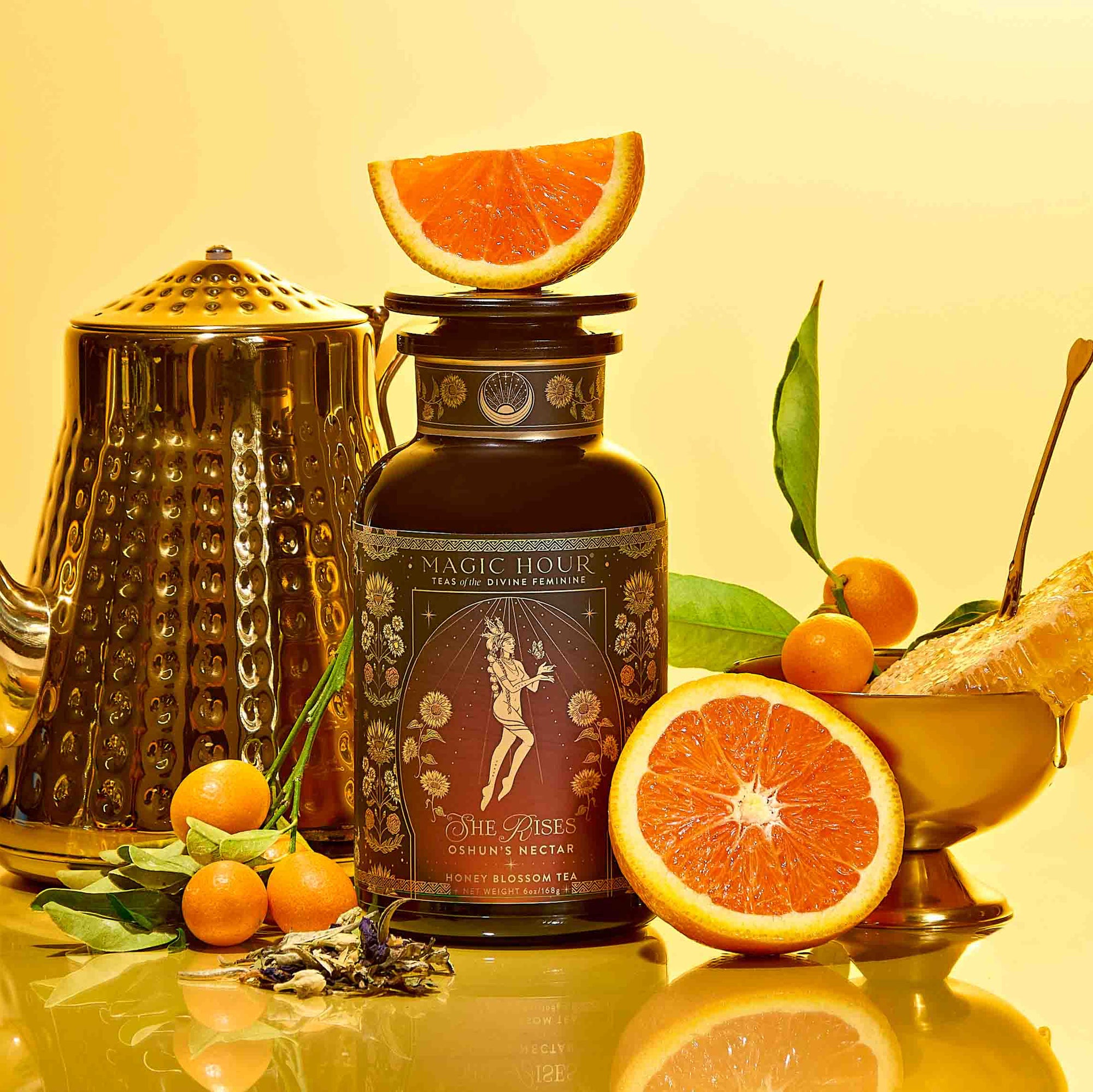 A tin of the Monthly Magic Tea Journey Subscription is surrounded by a gold teapot, orange slices, kumquats with leaves, loose tea, and a gold dish—all set against a yellow background—inviting you to discover the ritual guide inspired by goddess archetypes included with each enchanting tea subscription.