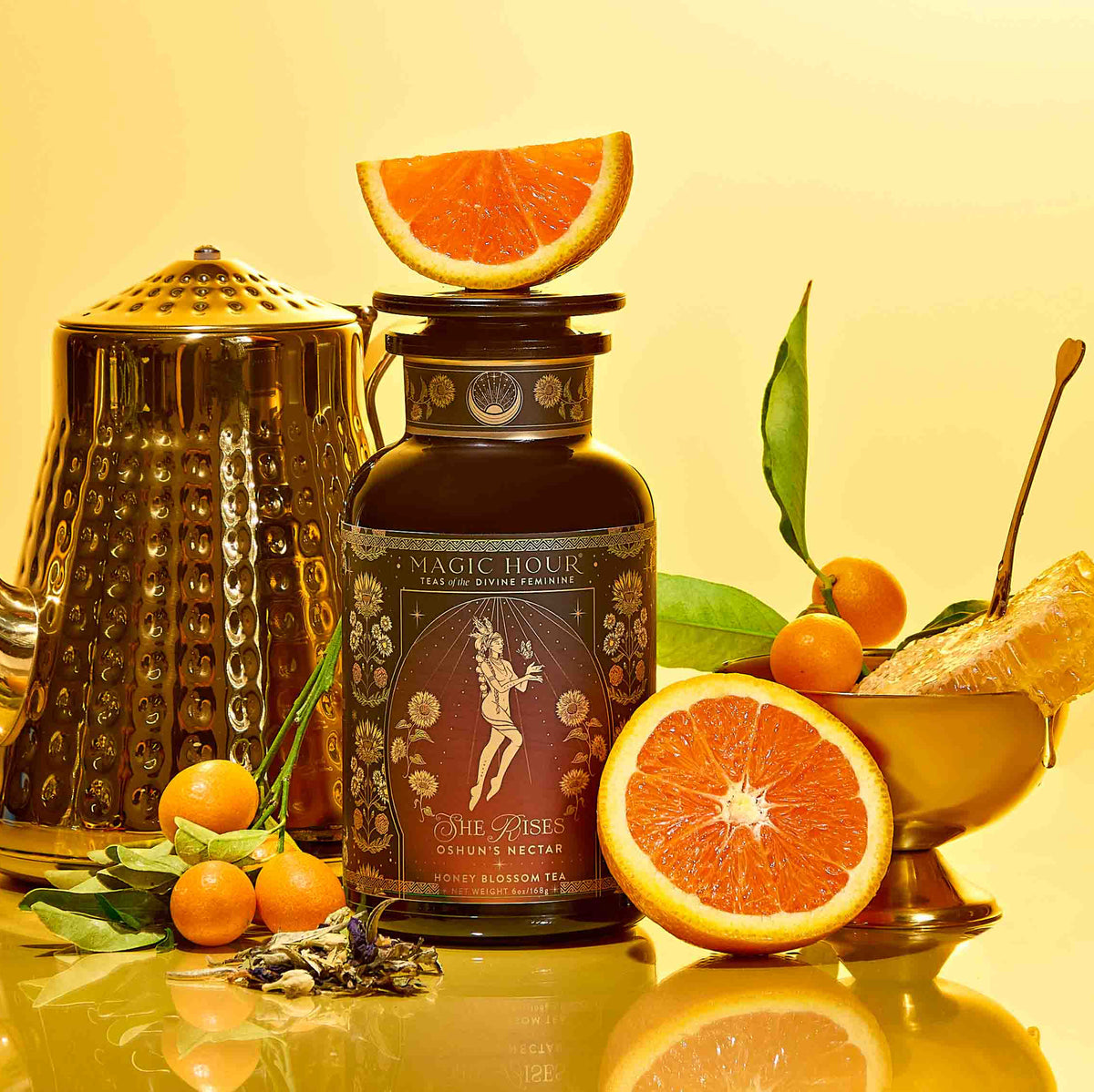 A gold teapot, a dark tea jar adorned with an illustrated label of Oshun’s Nectar: Honeyed Herbal Tea for Joy & Radiance, fresh oranges, kumquats, loose tea leaves, and a golden bowl with orange—all elegantly arranged on a yellow background—create an inviting tableau that captures the warmth and vibrancy of honeyed herbal tea.