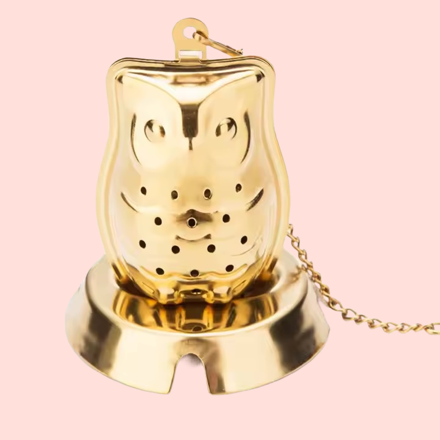 The Owl Strainer, an owl-shaped gold tea infuser featuring perforations and attached to a chain, rests on its matching gold base against a pale pink background.