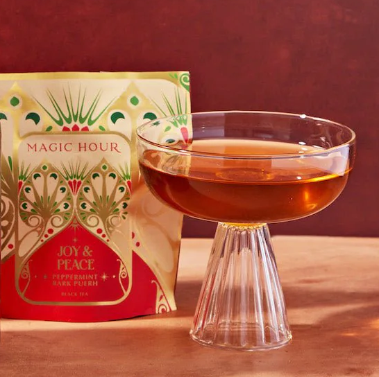 A glass cup filled with amber-colored liquid is placed on a wooden surface. Behind the cup is a decorative package labeled "Magic Hour" and "Joy & Peace" with "Peppermint Bark Pu-erh Black Tea." The festive design, part of The Best of Winter Box, features green and gold accents.