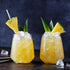 Two glasses of Pineapple Oolong cocktails are filled with ice, garnished with pineapple wedges and leaves. A straw rests in one glass, while extra ice is scattered on the dark surface, highlighting the vibrant tropical drink.