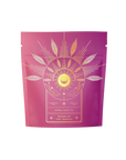 The packaging of this purple tea features gold floral designs with the text "Queen of the Tropics: Rosé all Day Alternative Tea." The design includes stylized leaves, a sunburst pattern, and subtle tropical fruit hints for an invigorating experience.