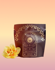 A sealed brown pouch labeled "Queen of the South: Delicious Cocoa Detox Tea - Magic Hour" with decorative gold designs, featuring a sun, crescent moon, and geometric patterns. Beside the Magic Hour pouch on a white surface lies a vibrant yellow flower.