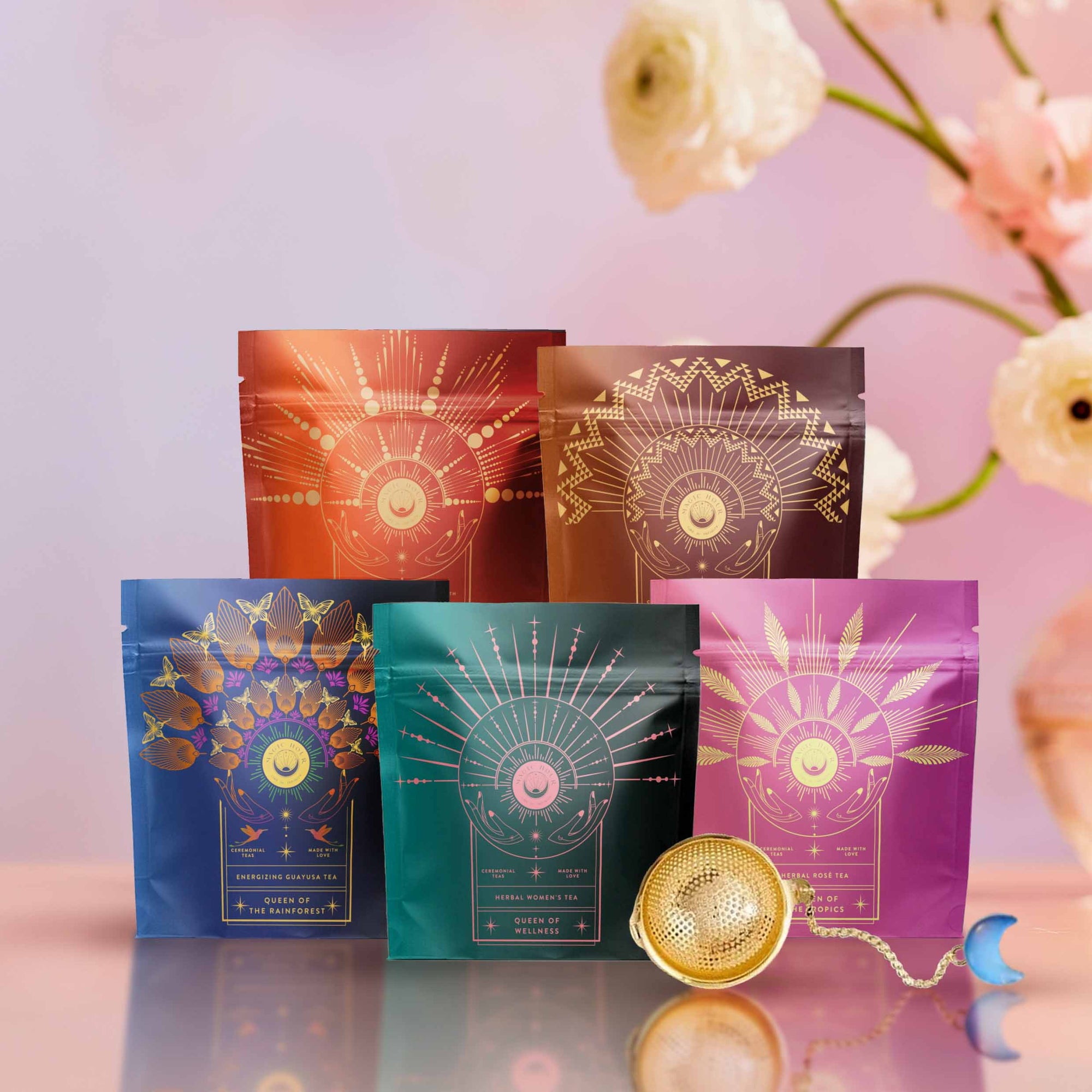 The Queen's Sampler Set features five vibrant tea packages with gold designs in a row. In front, a gold tea infuser and crescent moon charm complete this charming wellness tea set, accented by soft pink flowers on a pastel background.