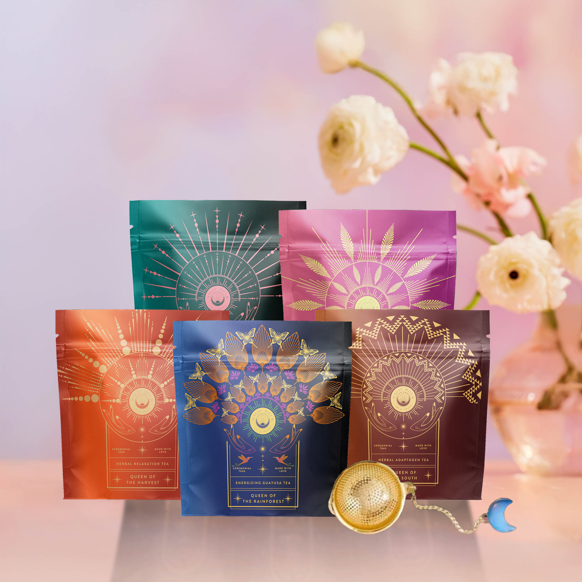 Five colorful tea packages from The Queen's Sampler Set, each with intricate gold designs, are arranged before a soft-focus background of white flowers. A gold tea infuser with a crescent moon charm completes the elegant gift set.