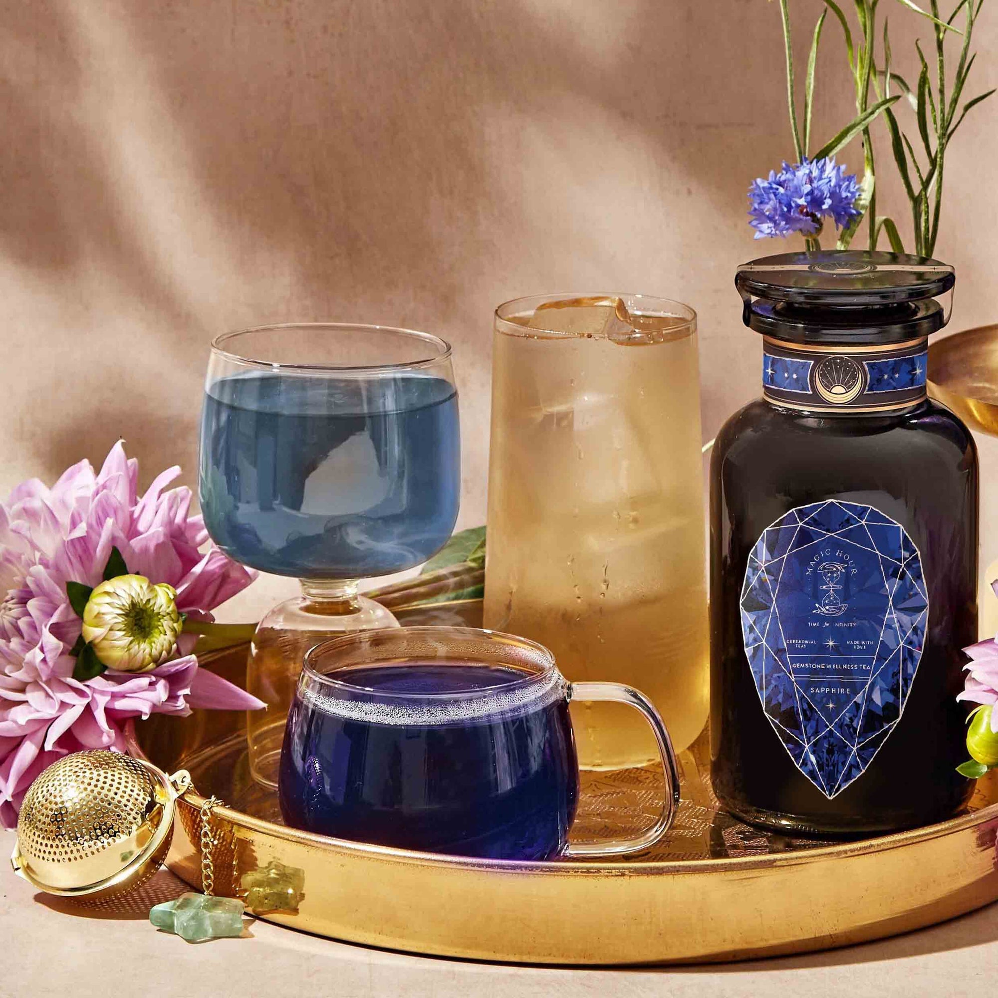 A gold tray holds a dark bottle, two bottles of Sapphire: Apple-Plum-Spice White Tea for Self-Expression, an iced drink, a tea infuser, and purple flowers. The background is warm beige with delicate green stems and a blue flower.
