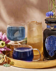 A gold tray holds a dark bottle, two bottles of Sapphire: Apple-Plum-Spice White Tea for Self-Expression, an iced drink, a tea infuser, and purple flowers. The background is warm beige with delicate green stems and a blue flower.