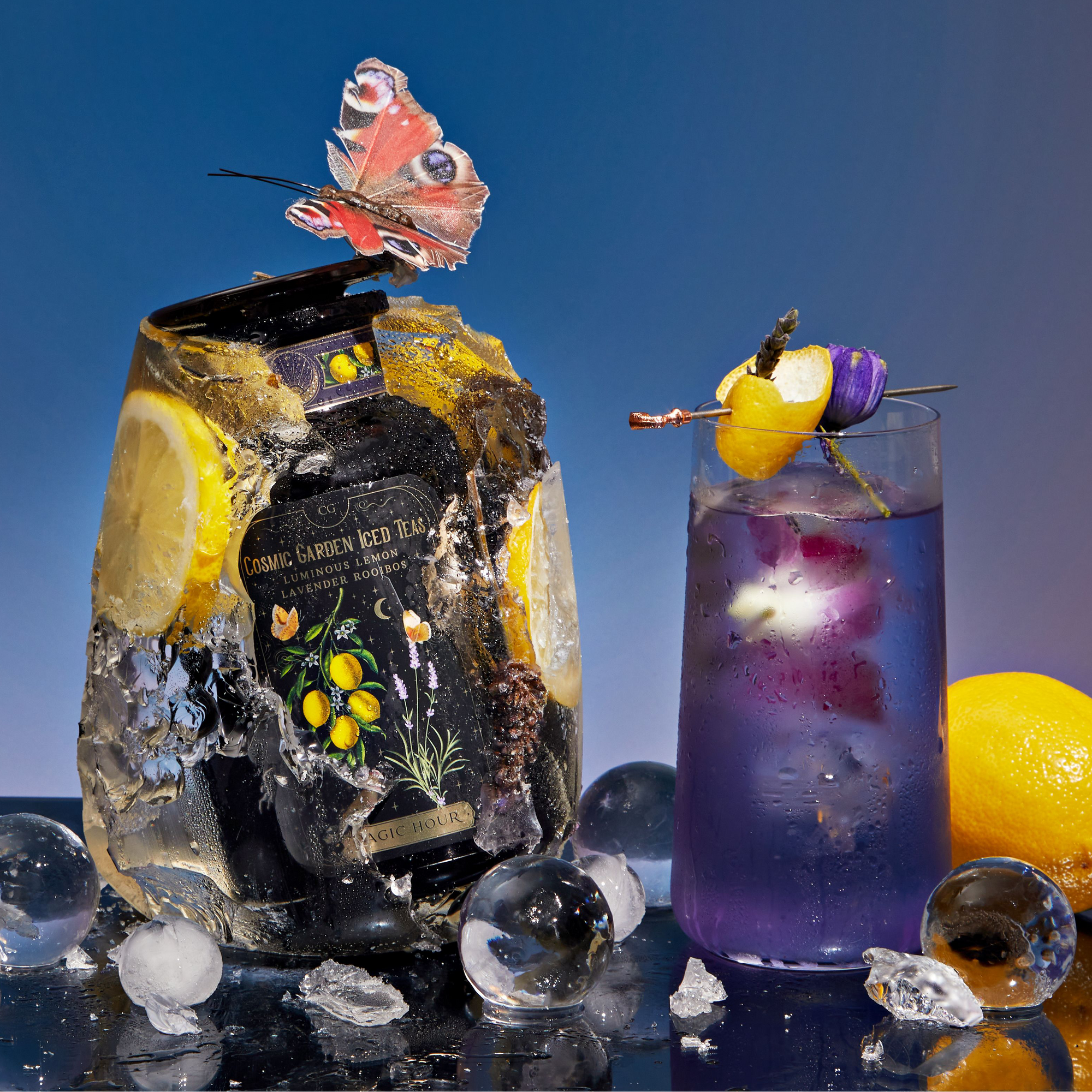 A bottle from the Cosmic Garden Iced Tea Sampler Set is encased in ice beside a purple lemon-garnished drink. A butterfly rests on the bottle, with teabags, ice spheres, and a whole lemon set against a blue gradient background.