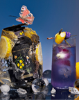 A bottle from the Cosmic Garden Iced Tea Sampler Set is encased in ice beside a purple lemon-garnished drink. A butterfly rests on the bottle, with teabags, ice spheres, and a whole lemon set against a blue gradient background.