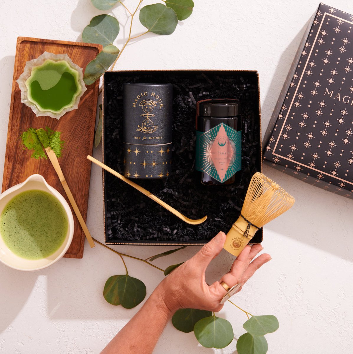 A gift set consisting of a black box with gold star patterns, including a jar of matcha powder, a wooden spoon, and a traditional bamboo whisk for making matcha. Next to the box are a cup of green matcha tea, a small dish of matcha powder, and eucalyptus leaves.