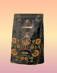 A dark amber bottle from Magic Hour, adorned with ornate label designs, features an illustration of a woman in a yellow dress surrounded by sunflowers and foliage. Titled "The Sun: Energizing Adaptogenic Herbal Tea with Yaupon, Moringa & Turmeric," the bottle has a black cap and displays intricate floral patterns with a mystical theme.