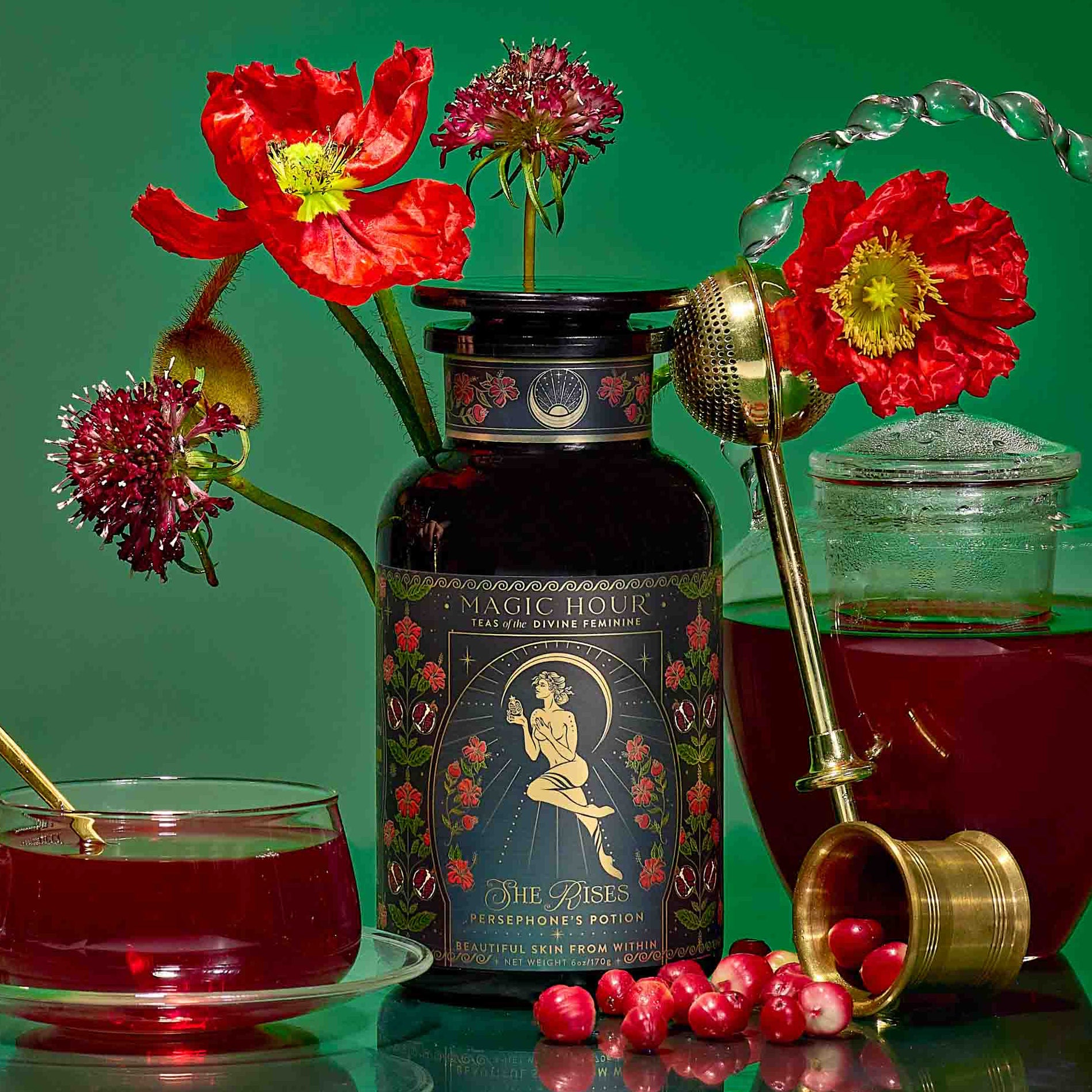 A tin of "Persephone's Potion: Pomegranate & Jasmine Green Tea" sits among red flowers, a glass teapot with pomegranate tea, a glass cup, a gold infuser, and cranberries on a vibrant green backdrop.