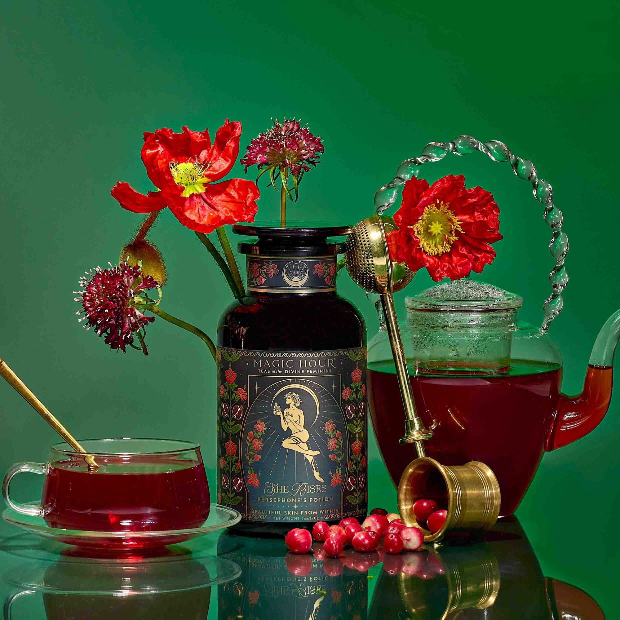 A tin of "Persephone's Potion: Pomegranate & Jasmine Green Tea" sits among red flowers, a glass teapot with pomegranate tea, a glass cup, a gold infuser, and cranberries on a vibrant green backdrop.