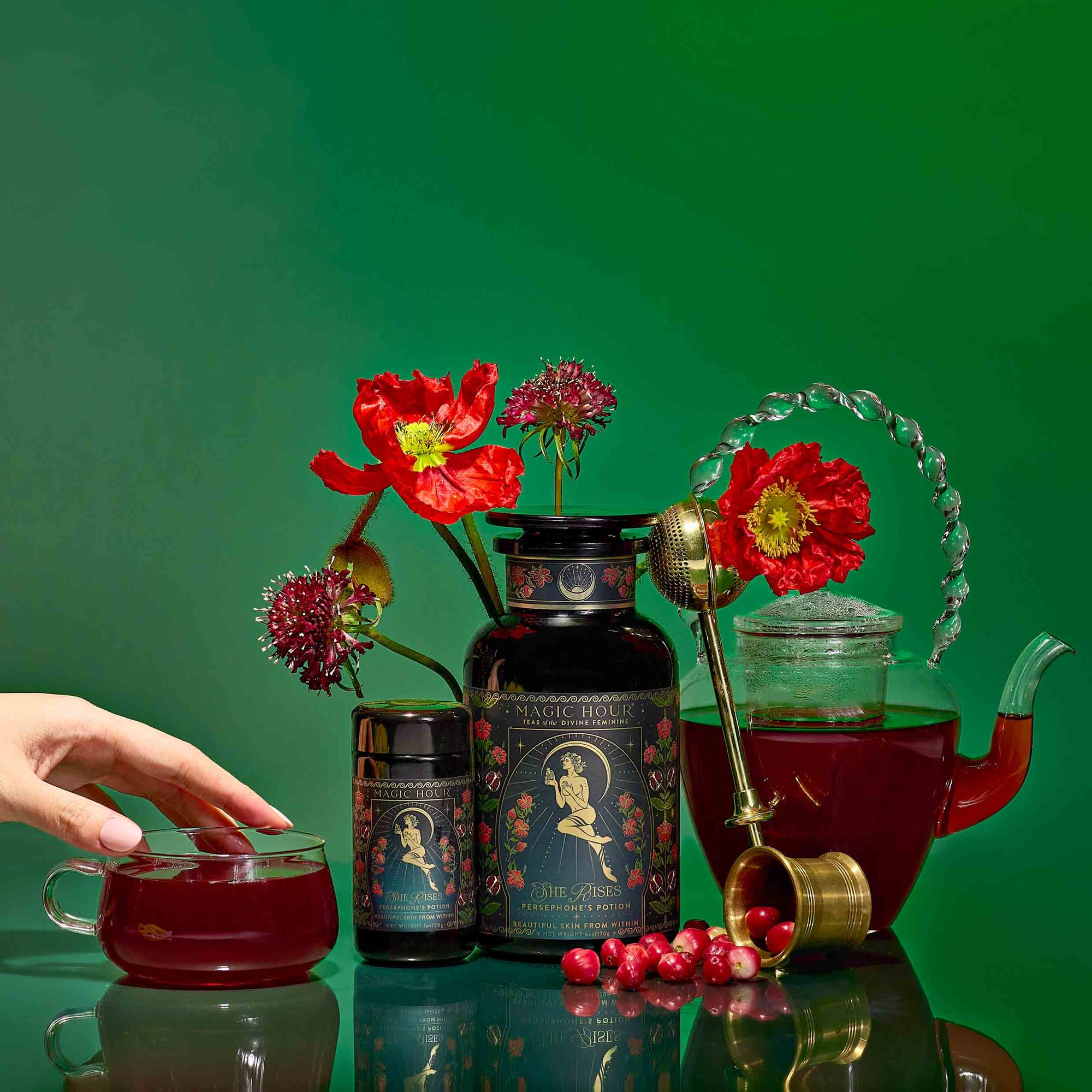 A hand reaches for a glass teacup filled with Persephone's Potion: Pomegranate &amp; Jasmine Green Tea, surrounded by ornate packaging, a teapot, red flowers, cranberries, and a tea strainer on a vibrant green background.