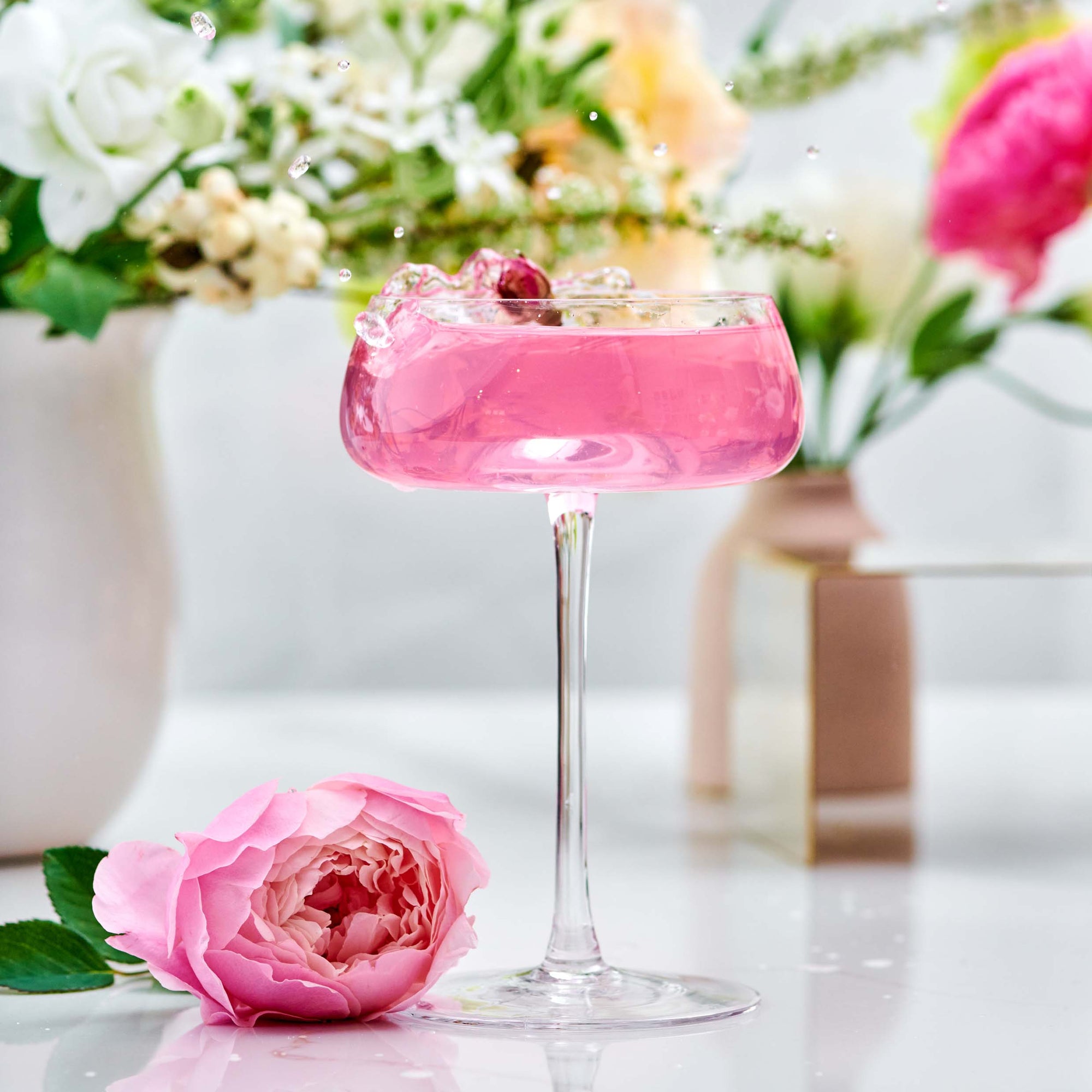 A pink cocktail in a coupe glass with a cherry, surrounded by splashes and a pink rose, is set against blurred vases of flowers. The scene evokes the bright elegance of Diamond - Champagne &amp; Strawberry Jasmine White Tea for Beautiful Skin.