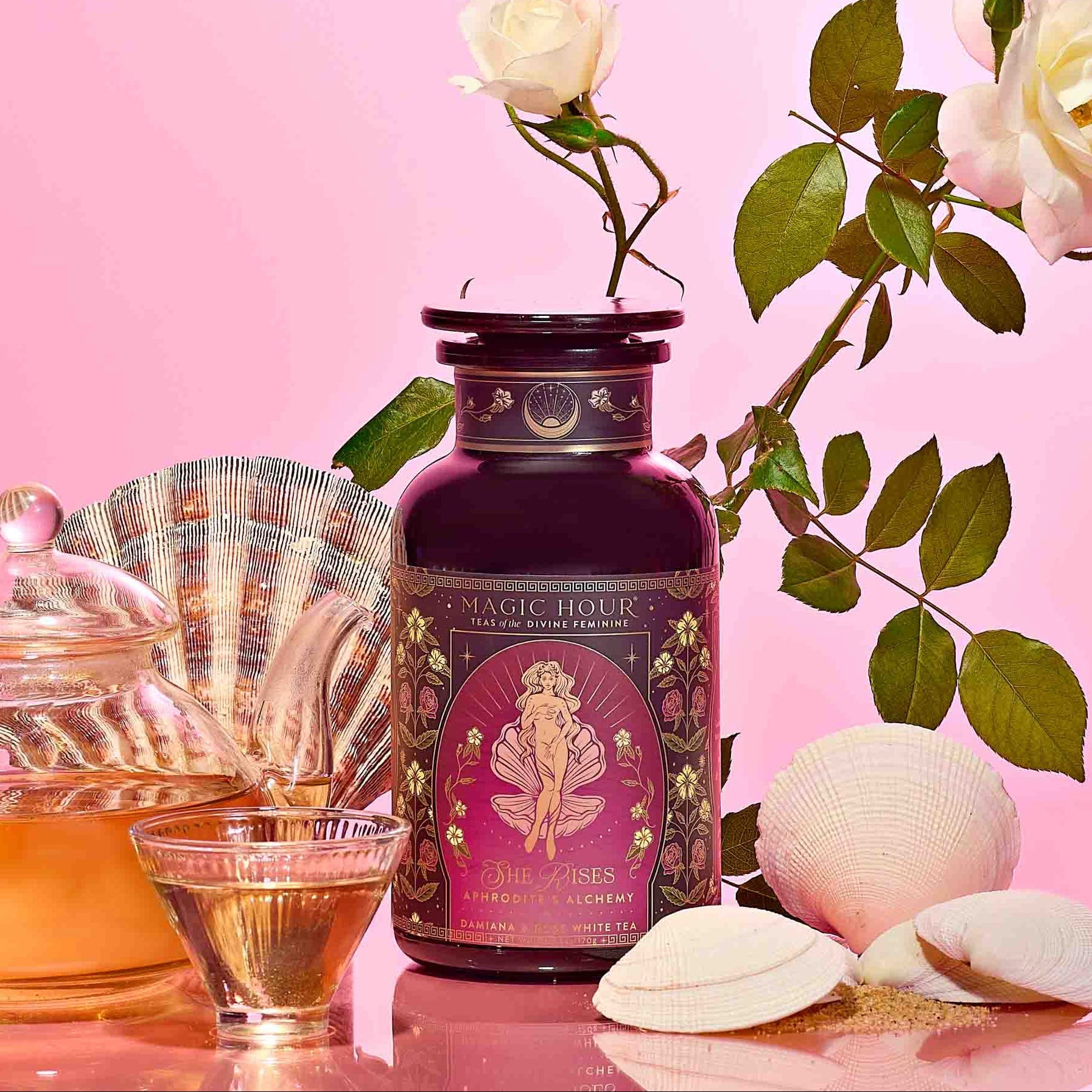 A glass teapot and cup of herbal tea accompany Aphrodite's Alchemy - Damiana & Rose White Tea, white seashells, and a white rose on a pink gradient background—an elegant scene inspired by the goddess of love.