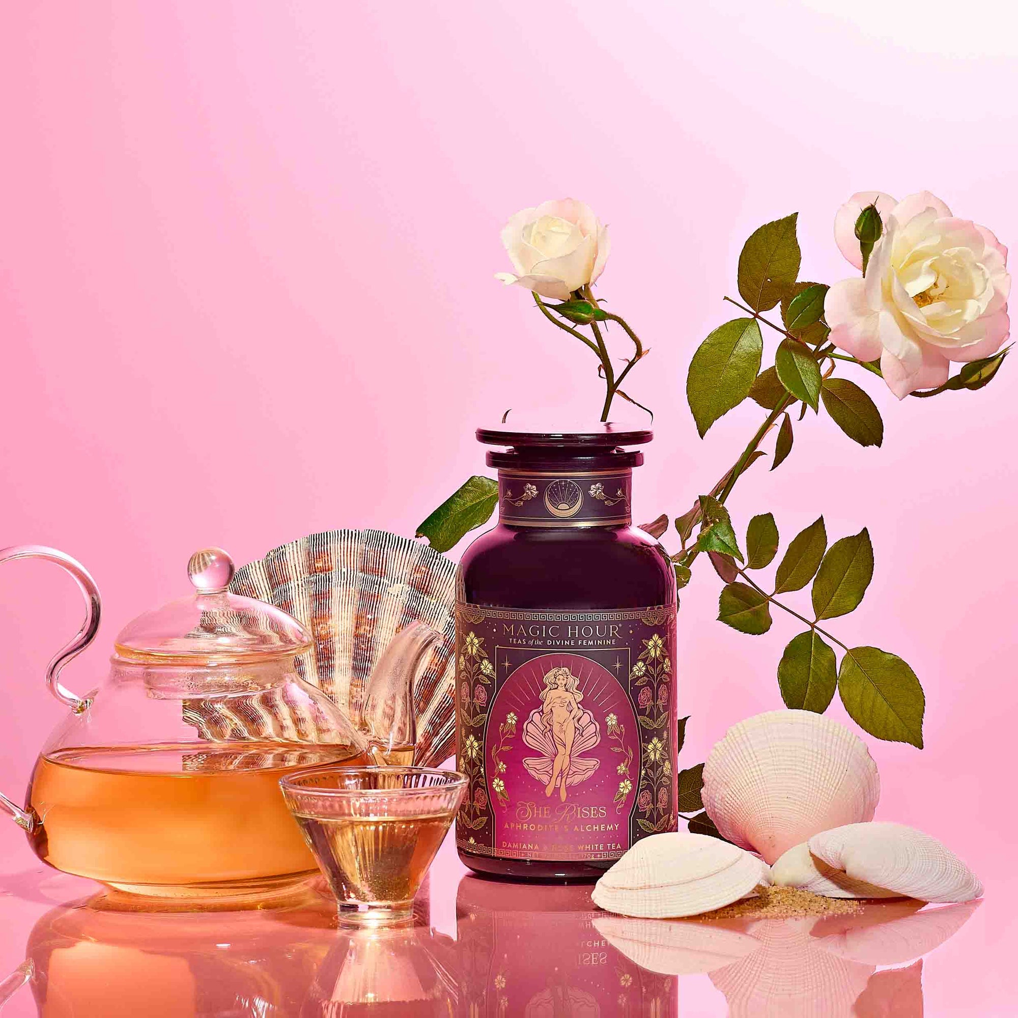 A glass teapot and cup of herbal tea accompany Aphrodite's Alchemy - Damiana & Rose White Tea, white seashells, and a white rose on a pink gradient background—an elegant scene inspired by the goddess of love.