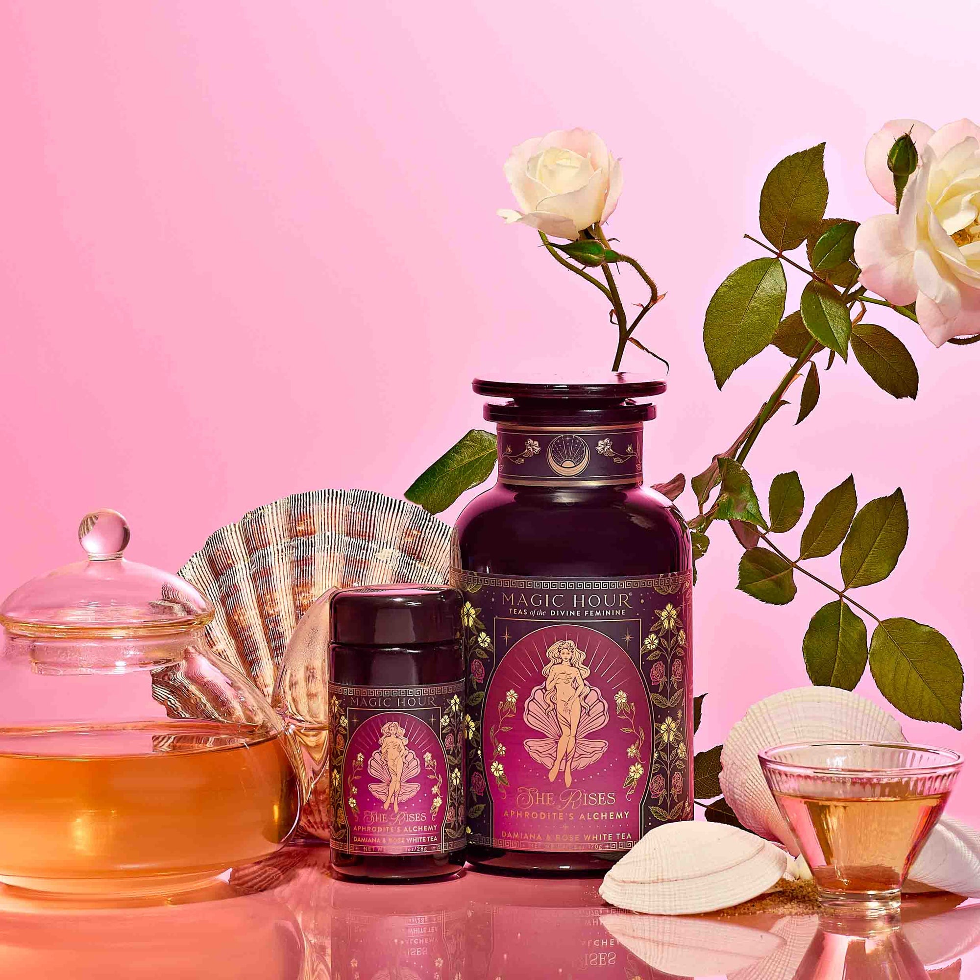 A glass teapot and teacup with tea, two ornate purple tins of Aphrodite's Alchemy - Damiana &amp; Rose White Tea, white roses, and seashells on a pink background evoke the elegance of Aphrodite, goddess of love.