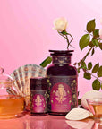 A glass teapot and teacup with tea, two ornate purple tins of Aphrodite's Alchemy - Damiana & Rose White Tea, white roses, and seashells on a pink background evoke the elegance of Aphrodite, goddess of love.