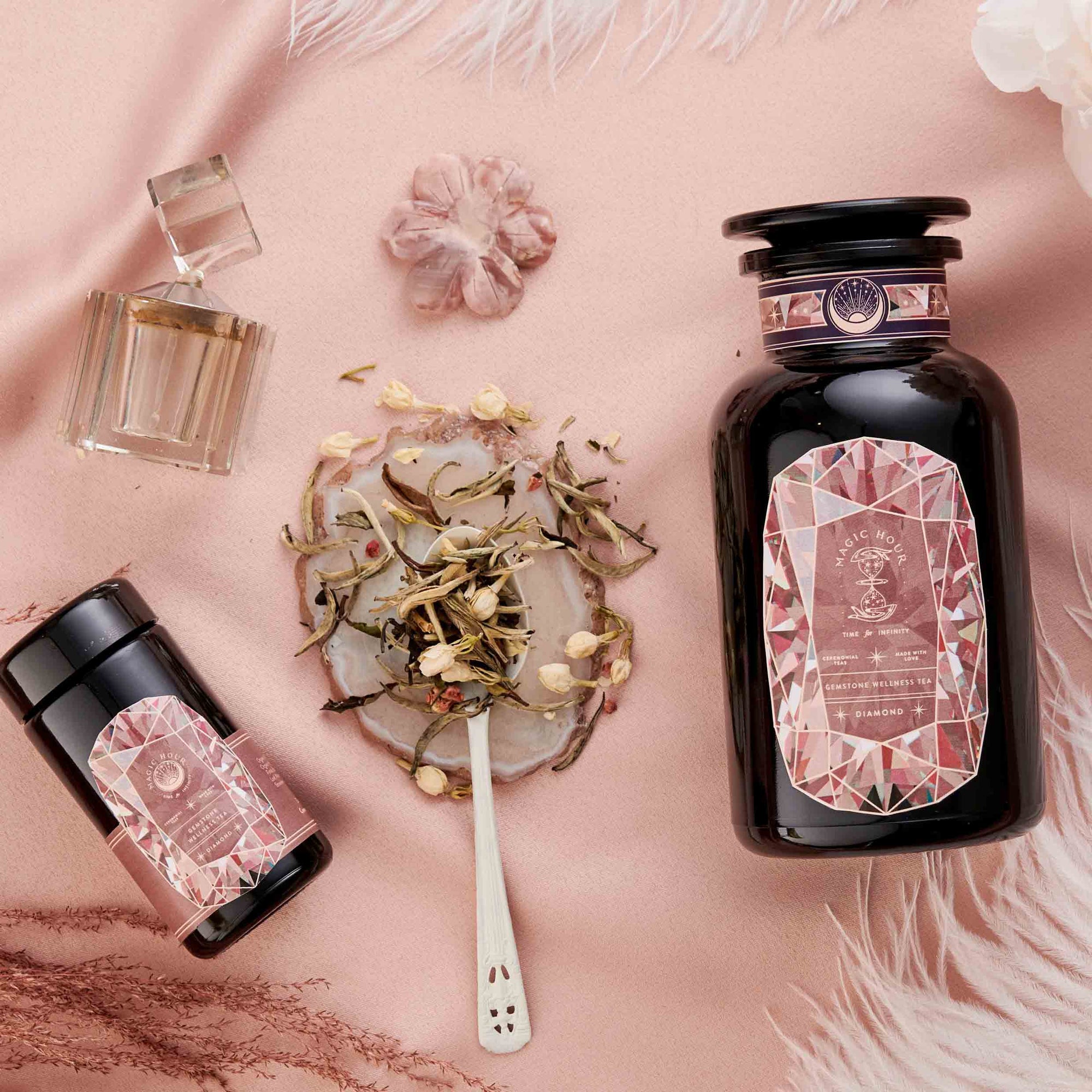 A flat lay on soft pink fabric features two Diamond - Champagne & Strawberry Jasmine White Tea jars with ornate pink labels, a clear perfume bottle, loose Silver Needle White Tea on a spoon, and scattered flower petals.