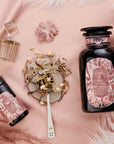 A flat lay on soft pink fabric features two Diamond - Champagne & Strawberry Jasmine White Tea jars with ornate pink labels, a clear perfume bottle, loose Silver Needle White Tea on a spoon, and scattered flower petals.