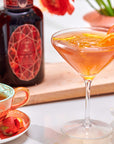 A martini glass with an orange cocktail and orange slices sits beside a decorative cup of Carnelian: Caffeine-Free Apricot Amaretto Tea and a dark bottle with a geometric label, flowers in the background.