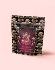 A pink, black, and gold tea package for the “Monthly Magic Tea Journey Subscription” is adorned with a floral design and an illustration of a four-armed woman in lotus pose, labeled “Magic Hour” and “She Rises: Lakshmi Abundance”—an invitation to a goddess ritual tea experience celebrating the Divine Feminine.