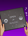 A person with blue painted nails and a ring on their right hand holds a black, rectangular box labeled "OPEN for MAGIC" with celestial illustrations. Two question marks float to the right of The Back to Realitea Mystery Box by Magic Hour, against a purple gradient background.