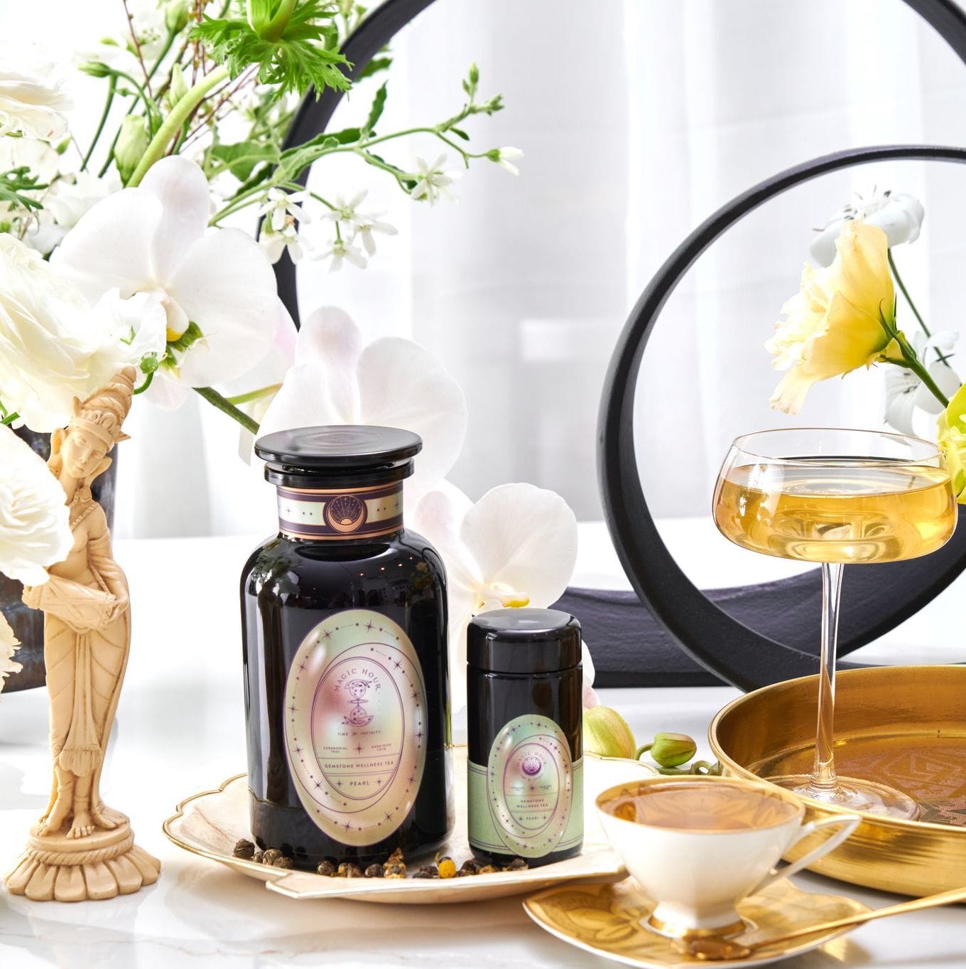 Elegant display featuring two black jars with pastel labels, a tall glass of golden liquid from Pearl Tea of Beauty, Clarity, and a Sense of Peace by Magic Hour, a gold saucer with a white cup, and delicate flowers in the background, including orchids. A small statue adds a touch of classical charm to the scene.