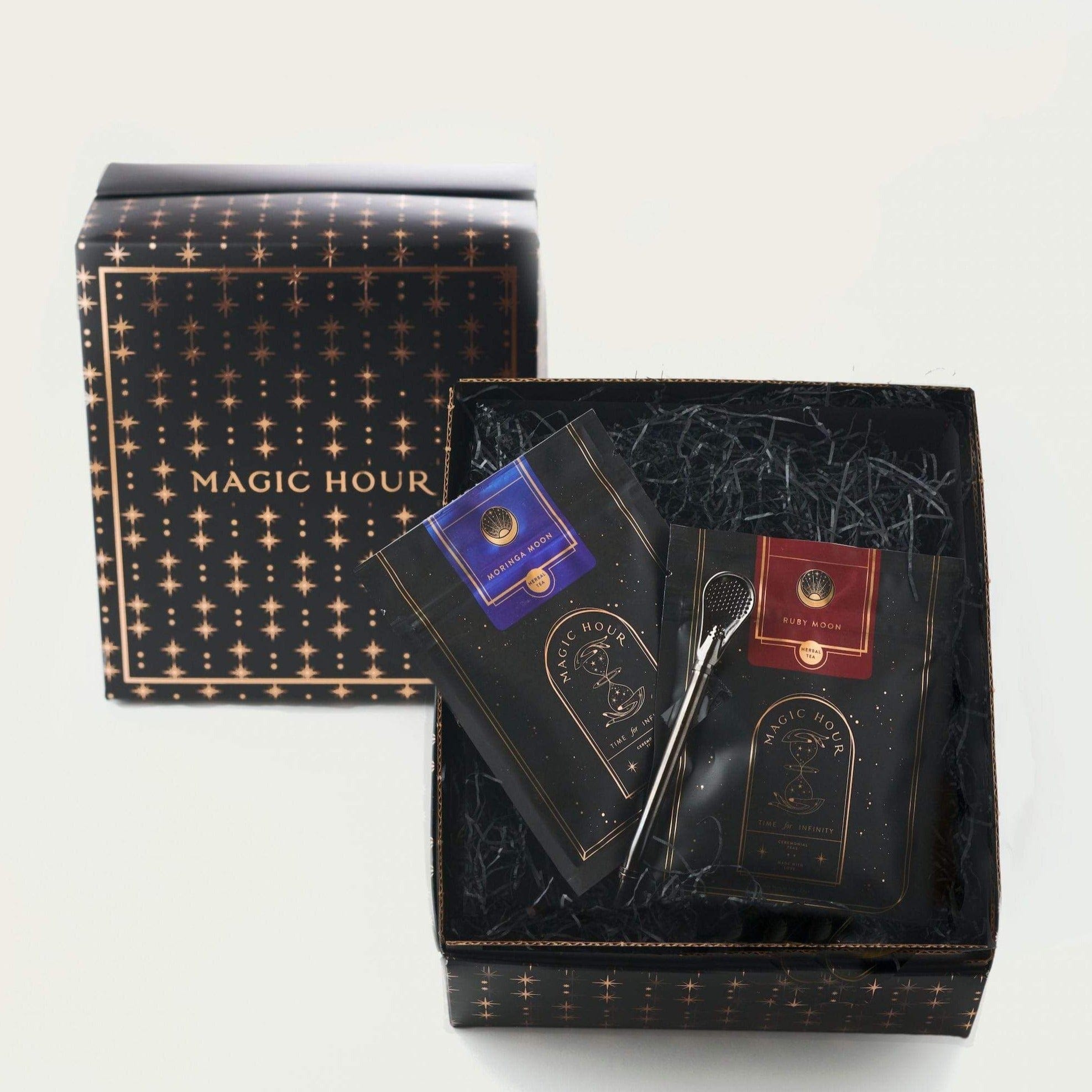 A gift box with a starry black design labeled "Magic Hour" on the lid contains two packets of luxurious loose leaf tea and a tea infuser. One packet is labeled "Blue Moon" and the other "Ruby Moon," both featuring elegant designs. The items are nestled in shredded black paper, making it the perfect Moon Lover: Herbal Immunitea Ritual Gift from Magic Hour.