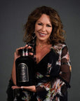 A woman with wavy brown hair smiles while holding a large, black bottle labeled "Big Magic: Violet Glass Apothecary Jar for Bulk Tea by Magic Hour." She is wearing a floral, patterned blouse or dress and standing against a dark gray background.