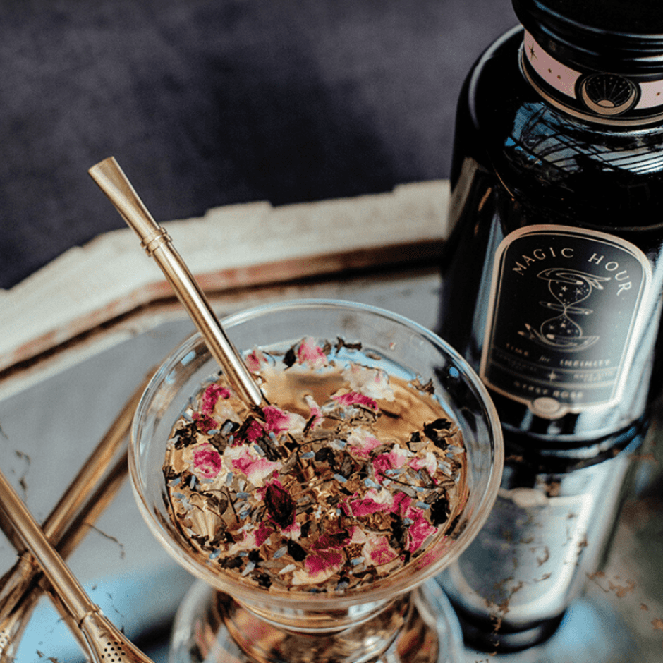 A jar labeled "Magic Hour" stands next to a glass filled with a mixture of herbs and dried rose petals, complemented by an Alibaba Sips Tea Straw &amp; Strainer Bombilla resting inside. Both items are set on a reflective surface, beautifully capturing the light.