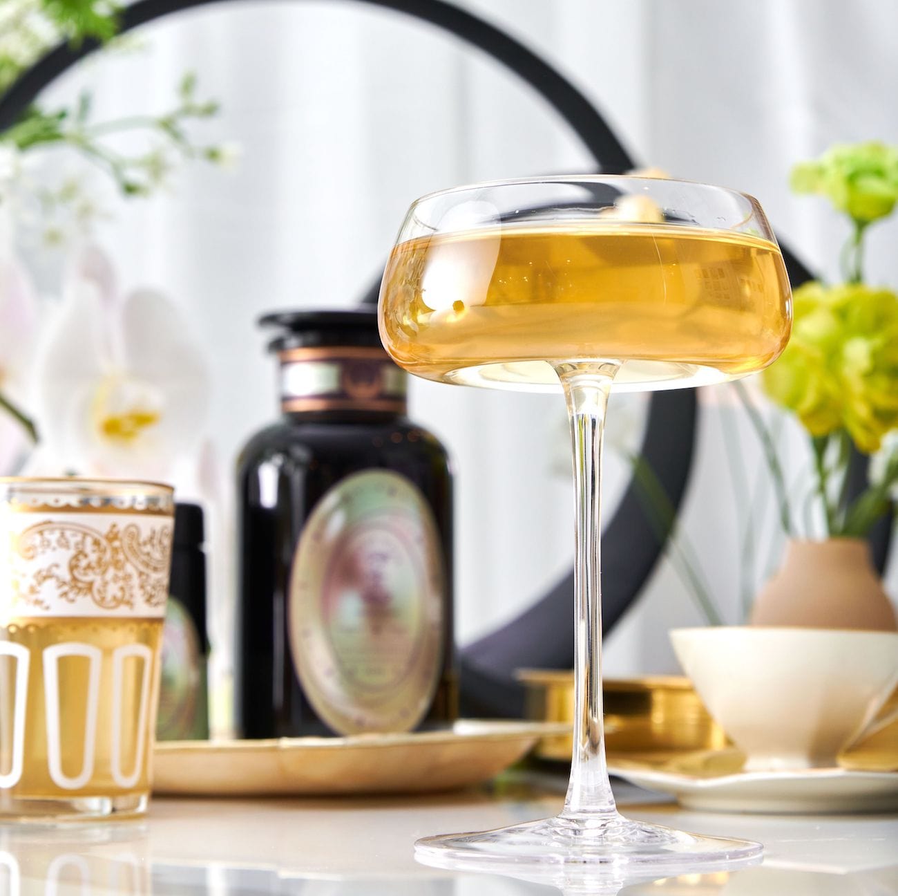 A clear, elegant coupe glass filled with a golden beverage sits in the foreground on a white table with an ornate white and gold glass to its left. The background features a blurred black circular frame, white flowers, and a decorative black apothecary jar hinting at the charm of Magic Hour's Pearl Tea of Beauty, Clarity, and a Sense of Peace.