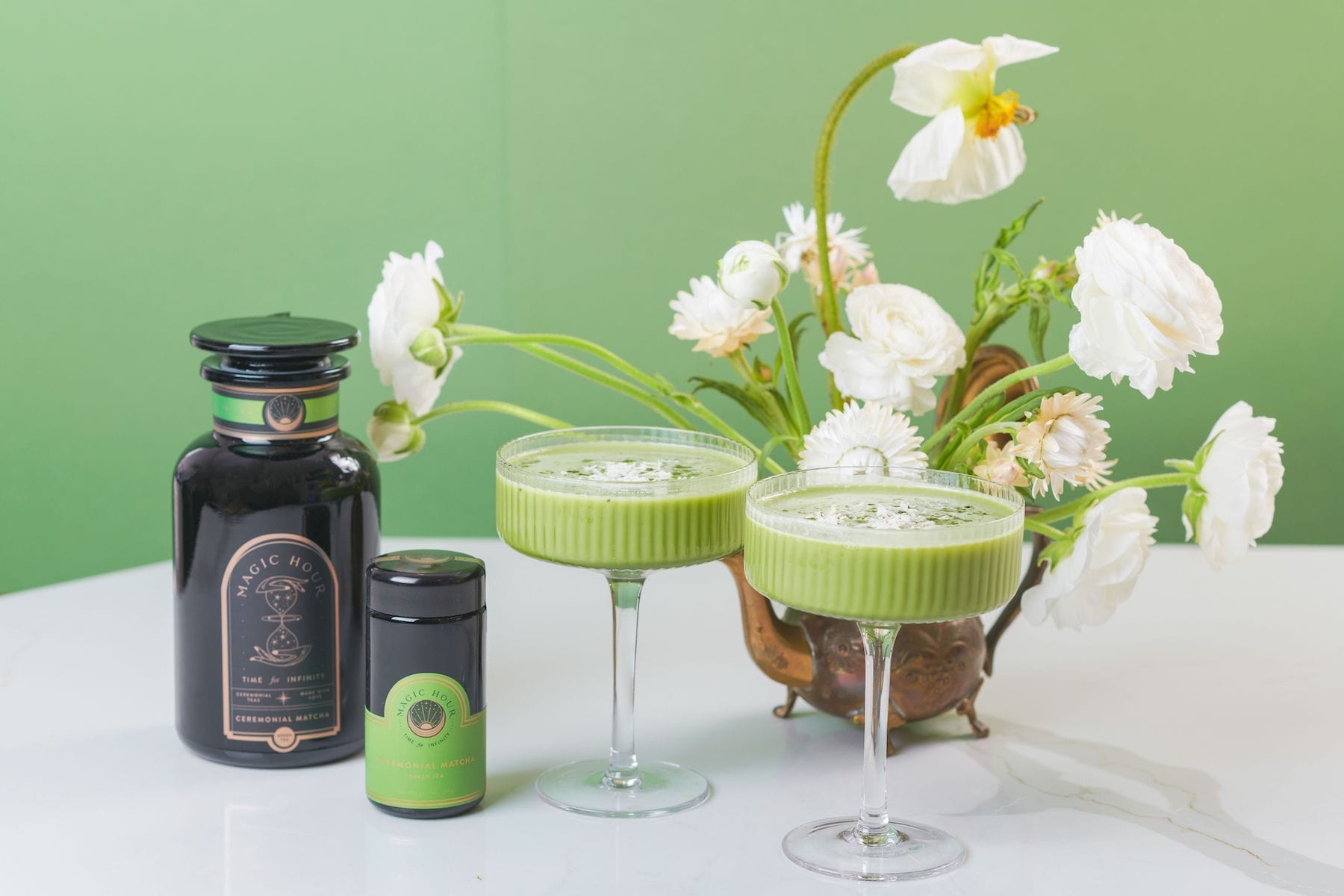 Two glasses of a green matcha drink are placed on a white surface. Beside them are two containers of organic matcha powder and syrup, with elegant black and green packaging. In the background, there is a decorative vase filled with white flowers against a green wall, embodying the essence of Magic Hour's Ceremonial Matcha Traveler Gift Box.