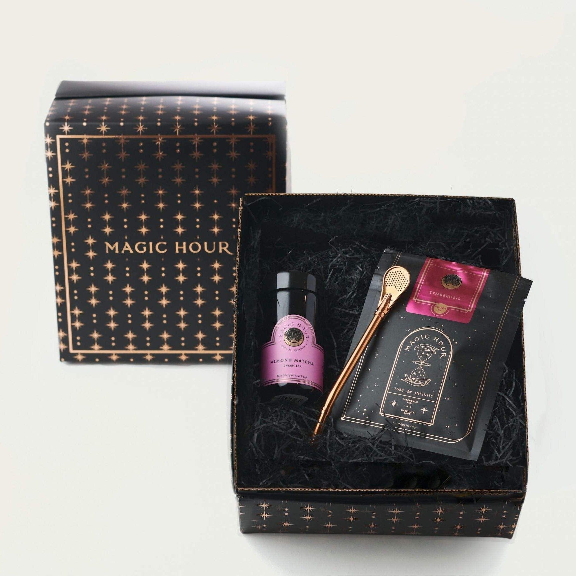 A luxury gift box labeled "Magic Hour Sip Wellness: Ritual Kit for Immunity & Joy" with a constellation design. Inside is a bottle with a pink label, a golden stirring spoon, a packet of organic tea, and a booklet. The box has black shredded paper as padding. The lid is placed leaning against the box.
