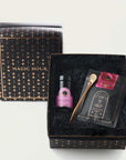 A luxury gift box labeled "Magic Hour Sip Wellness: Ritual Kit for Immunity & Joy" with a constellation design. Inside is a bottle with a pink label, a golden stirring spoon, a packet of organic tea, and a booklet. The box has black shredded paper as padding. The lid is placed leaning against the box.