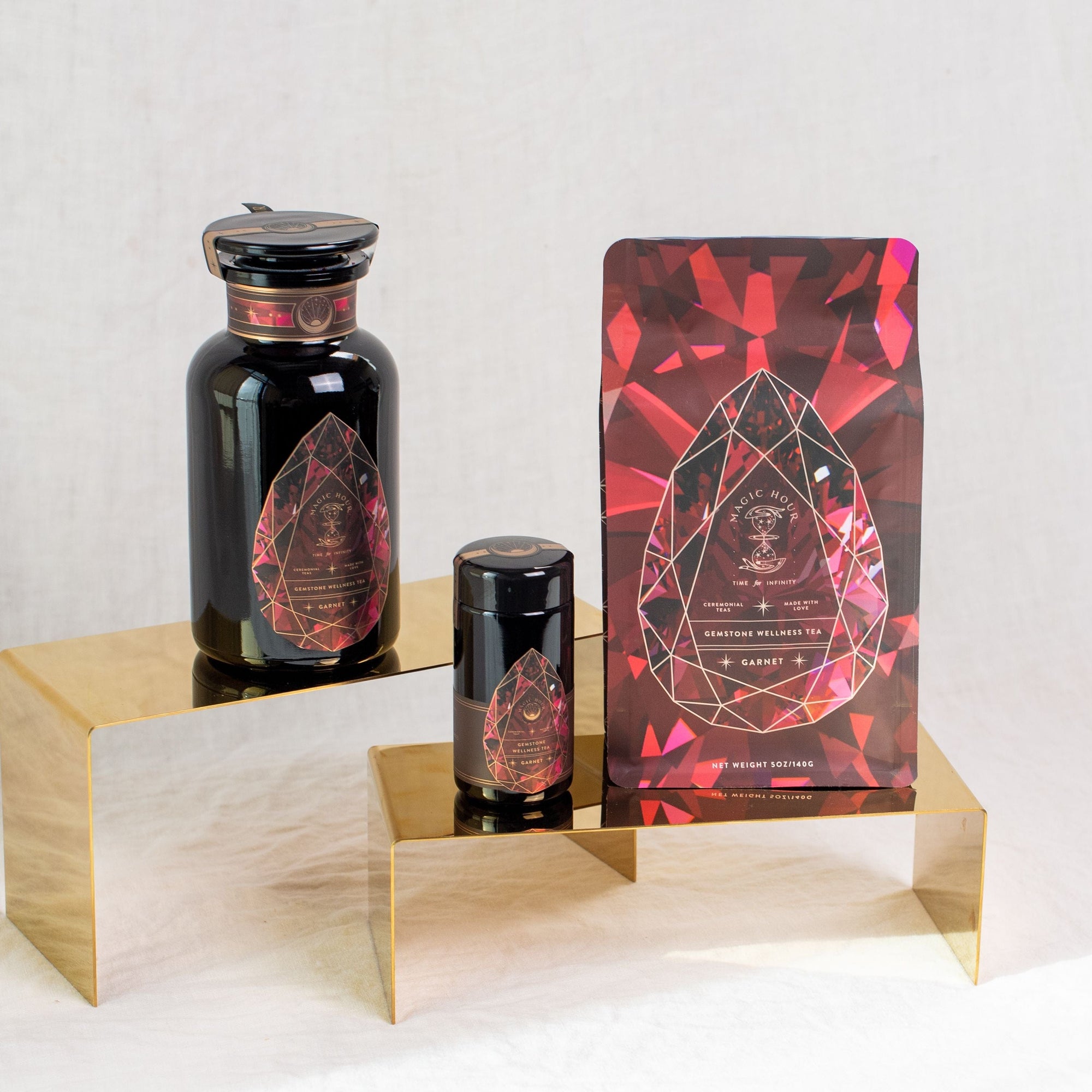 Three luxurious dark-hued containers with intricate red and gold geometric designs, labeled as Magic Hour's Gemstone Bundle: Garnet, Amethyst, Aquamarine, are elegantly displayed on golden stands against a light background. The products include a large bottle, a smaller jar, and a resealable bag.
