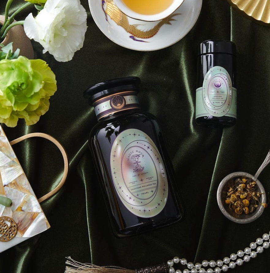A luxurious flat lay featuring a tea set with a cup of Pearl Tea of Beauty, Clarity, and a Sense of Peace by Magic Hour, a black tea container, a small jar, a bowl of loose leaf tea with a spoon, white and green flowers, a string of pearls, a folded fan, and decorative items on a dark green velvet background.