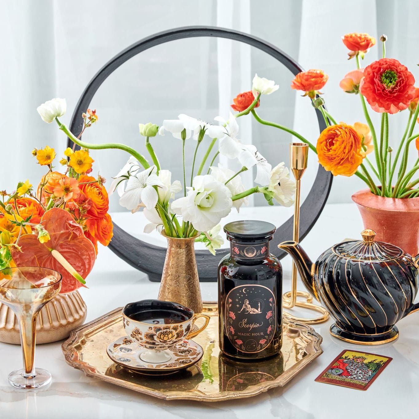 A decorative setup featuring a Magic Hour Scorpio Tea for Sensual Brilliance tin, vintage teacup and saucer, black teapot, vases with white and orange flowers, and a gold-trimmed tray. Additional items include a gold watering can, a small round mirror, and a wine glass, all set on a white surface.