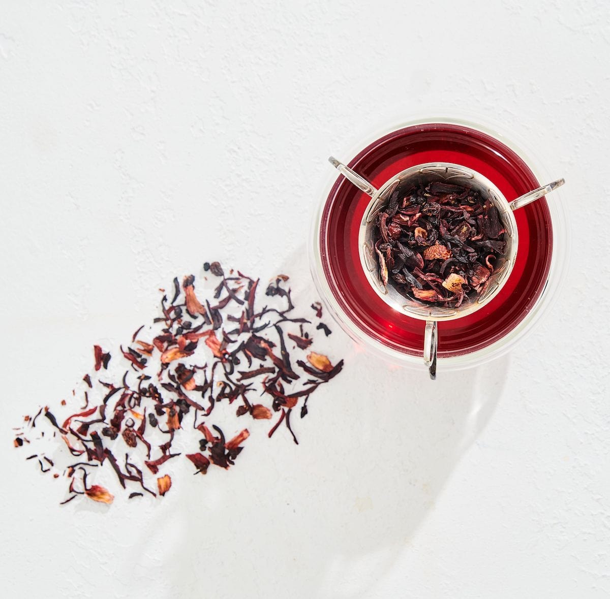 A glass cup filled with vibrant Magic Hour Immune Boosting Teas Sampler Set sits on a white surface. Loose leaf tea and petals are scattered beside the cup, while a metal tea infuser containing organic tea is placed on top, casting a delicate shadow.