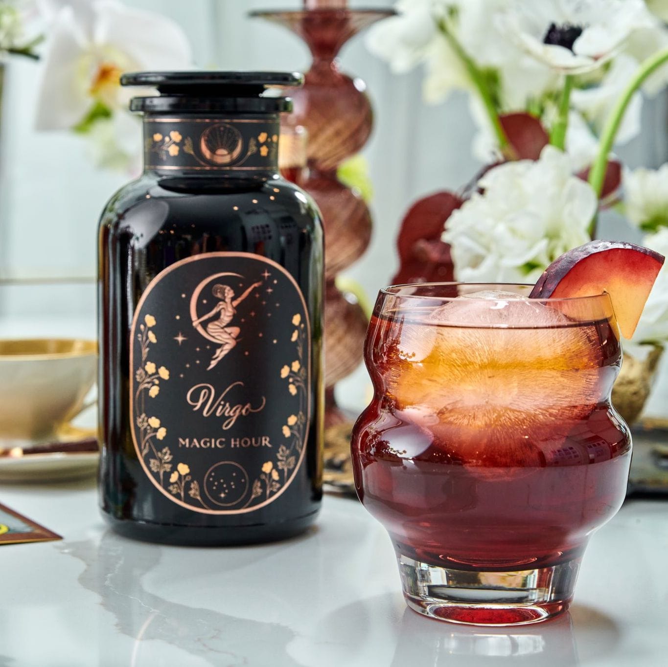 A glass of iced plum-colored drink garnished with a plum slice sits next to a black bottle labeled &quot;Virgo Tea of Virtue, Wit &amp; Meticulous Magic&quot; decorated with gold celestial designs. The background features a teacup, white flowers, and decorative vases, evoking the soothing essence of Magic Hour.