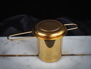 A gold-colored Luxury Traveler Gift Box from Magic Hour, designed for loose leaf tea, boasting a cylindrical mesh body and two extended handles, rests elegantly on a white marble slab. The strainer's lid matches in gold color, and the black background adds a touch of sophistication.