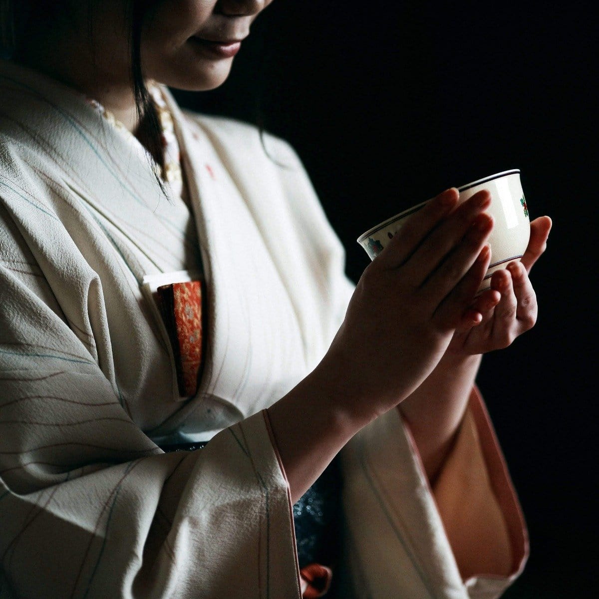 A person dressed in a traditional Japanese kimono is holding a ceramic bowl of Kansai-Kyoto Ceremonial Matcha by Magic Hour with both hands. The image is dimly lit, focusing on the serene and delicate gesture, with the individual’s face partially visible.