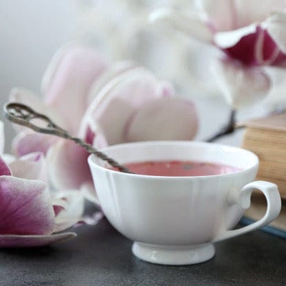 A white teacup filled with pink liquid, placed next to a silver spoon. Pink and white magnolia flowers and a book are also visible in the background, creating a serene and delicate ambiance—a perfect setting for enjoying Magic Hour Magnolia Rose Oolong Tea.