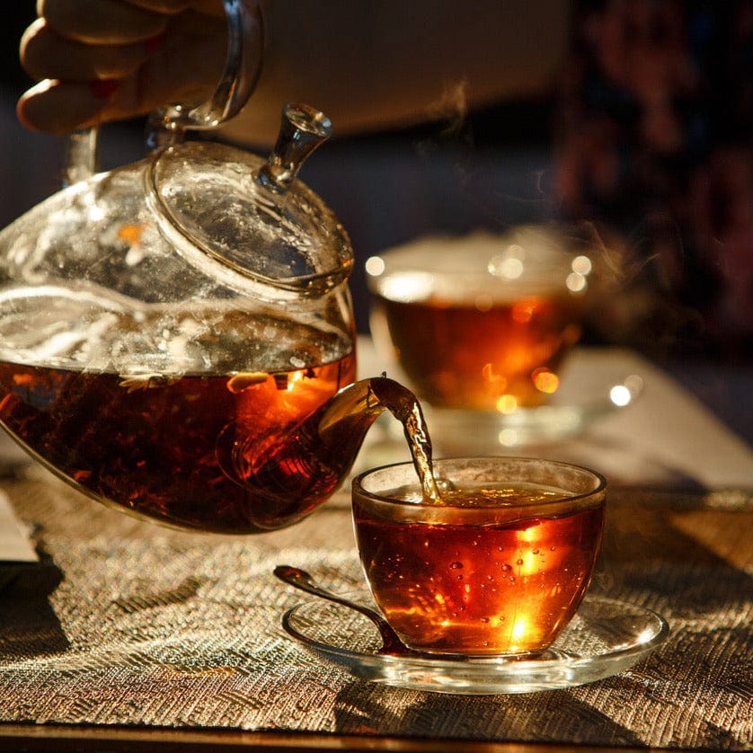 A hand pours steaming Tea of The Rising Sun : Japanese Breakfast from a clear glass teapot into a matching glass teacup. Sunlight highlights the amber color of the Magic Hour loose leaf tea. Another filled teacup is blurred in the background, set on a table with a woven placemat.