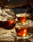 A hand pours steaming Tea of The Rising Sun : Japanese Breakfast from a clear glass teapot into a matching glass teacup. Sunlight highlights the amber color of the Magic Hour loose leaf tea. Another filled teacup is blurred in the background, set on a table with a woven placemat.