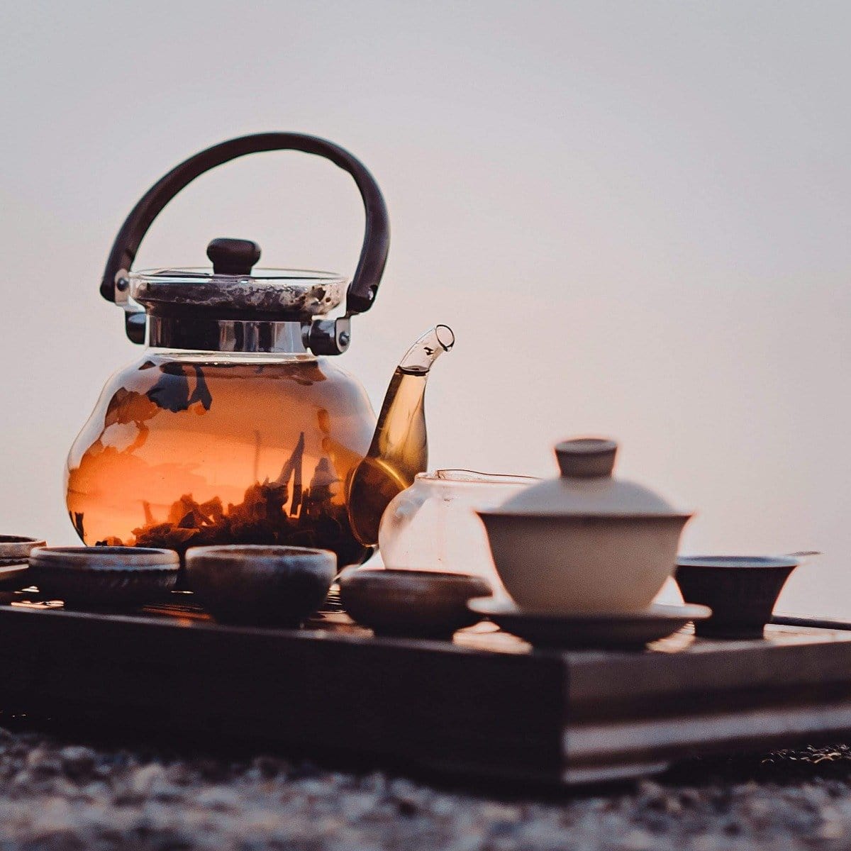 A glass teapot filled with Sin Eraser™ : Puerh Tea from Magic Hour sits on a wooden tray alongside several small cups and bowls. The tea has a warm amber hue, and the setup is outdoors, suggesting a serene, peaceful atmosphere.
